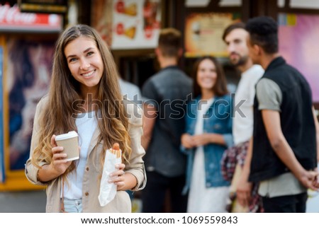 Happy girl enjoying street food, holding a cup of cappuccino and a hotdog. Fast food kiosk in the background.