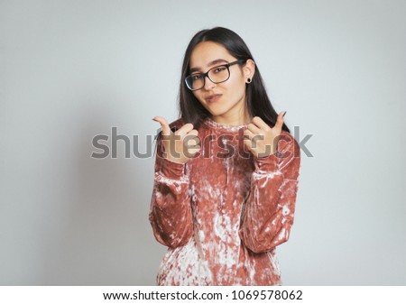 beautiful asian woman shows a sign she likes, thumbs up, wears glasses and pink sweater, studio photo on background
