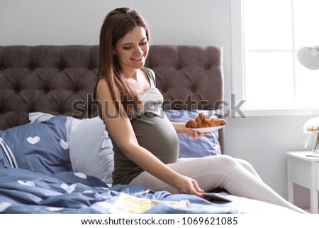 Young pregnant woman holding plate with croissant on bed