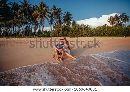Happy romantic couple sitting on tropical beach on palm trees background, hugging each other and enjoy sunset. Vacation, travel concept. Honeymoon