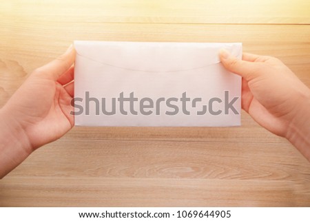 Woman's hand holds a white long paper envelope or letter envelopes on a wooden desk with natural sunlight in morning, Top view background.