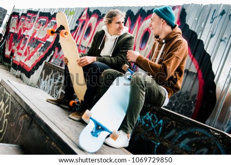 Skateboard is passion. Two young men talking to each other with smile while sitting in skateboard park with longboards outdoors 