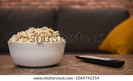 White bowl of white salty popcorn and remote control on a wooden table in front of a couch. Front view, blurry background
