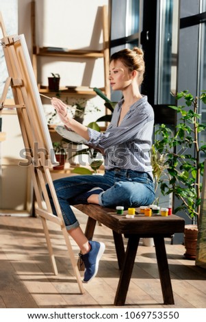 side view of female artist sitting on bench with paints and drawing picture on easel 