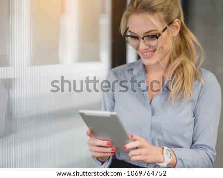 Cheerful young woman is watching something on tablet with interest. She is standing in formal clothing and smiling. Copy space