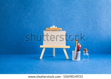Wooden easel with textured blank paper canvas on blue background. Beautiful art class studio interior, watercolor brushes, pencils in a case, water. Artist's advertising poster mockup