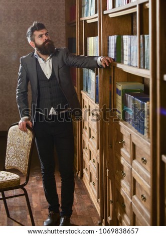 Intellectual elite concept. Aristocrat on busy face looking for book. Man in classic suit stand in vintage interior, library, book shelves on background. Oldfashioned man looking at books, seeking.