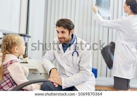 Male doctor talking with little patient while his colleague working in the background