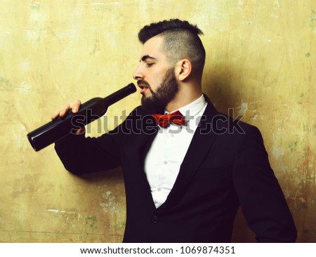 Young classy man with tired face expression drinks alcoholic beverage out of long glass bottle, on old beige wall background. Concept of Friday night and alcohol