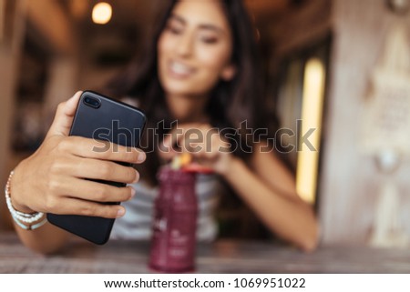 Woman taking a selfie with a smoothie placed on the table using a mobile phone for her food blog. Food blogger shooting photos for her blog at home.