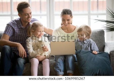 Happy family with adopted children have fun using laptop sitting on sofa at home, parents and son daughter relaxing on couch holding computer, smiling man woman with kids watching funny video online
