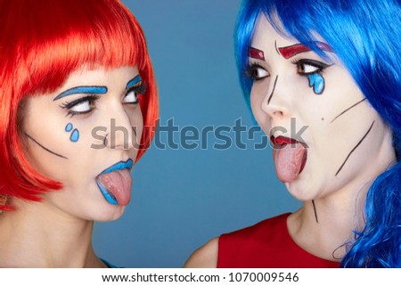 Portrait of young women in comic pop art make-up style. Females in red and blue wigs on blue background. Girls show each other tongues