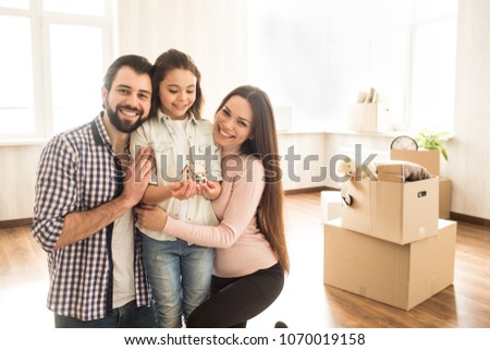 A picture of father, mother and their child standing in a bright room. Parents are hugging their child and smiling while girl is holding a wood toy house and looking at it.