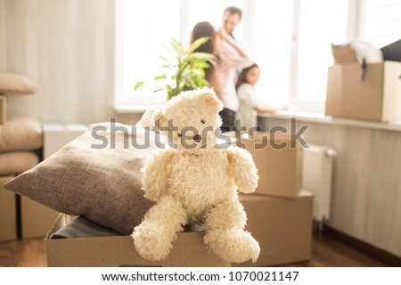 Cute picture of white toy of bear sitting on the box with pillows. He is looking straight ahead. Also there is a family in the room. They are looking outside from the window and talking.