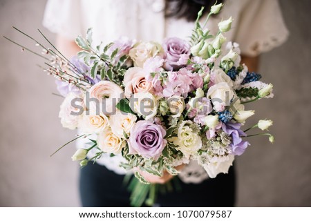 Very nice young woman in a white shirt holding blossoming flower bouquet of  fresh ranunculus, roses, carnations, eustoma in cream and purple colors on the grey wall background Royalty-Free Stock Photo #1070079587