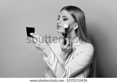 Young pretty woman in blue suit holding makeup brush and small mirror and applying cosmetics. Make-up artist. Girl with long straight blonde woman posing against pink background