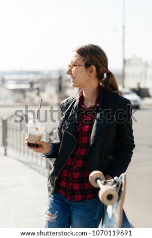 Blonde woman wearing black jacket with cup of coffee and her longboard in the city center in sunset sunshine