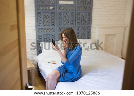 Beautiful girl in blue dress is sitting on white bed linen in minimalist loft design room. Young woman is doing make up. She is looking in the mirror and powders her face.