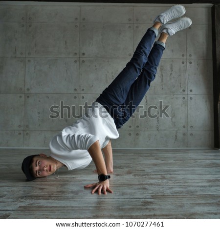 Teenage boy in white sweater and baseball cap, jeans and sneakers makes breakdance in dance studio. Dynamics of modern dance movement. He makes a stand upside down