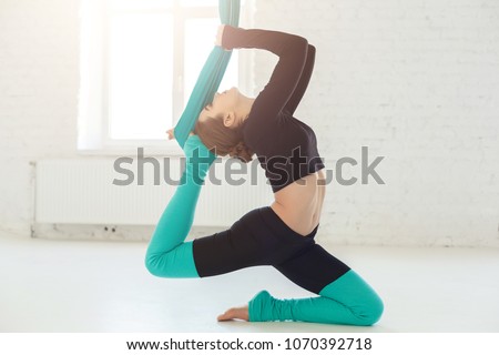 Young slim woman practicing fly yoga asana over white window background in fitness studio, copy space. Health, sport, yoga concept
