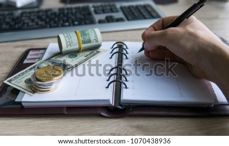 Bitcoin crypto currency on top dollar biils on notepad with mans hand with pen. Profit from mining crypto currencies. Miner with dollars from trading. Record of money income in notebook from mining.