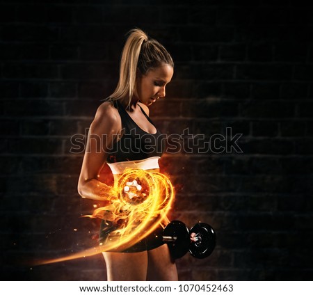 Attractive young blond woman doing bodybuilding, holding fire dumbbells. Concept of hard work and motivation, dark brick wall background. Very high resolution image Royalty-Free Stock Photo #1070452463