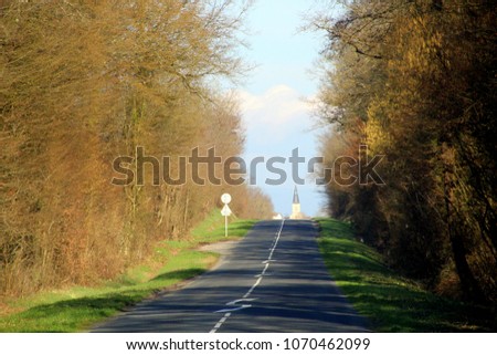 Magic forest road: an asphalt covering with white markings between two rows of spring trees with green young leaves, spire of small Catholic French church on horizon and blue sky with clouds