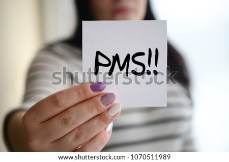 Young sad girl shows a white sticker. Caucasian brunette holding a sheet of paper with message. PMS  Royalty-Free Stock Photo #1070511989