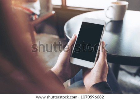 Mockup image of woman's hands holding white mobile phone with blank black desktop screen in cafe