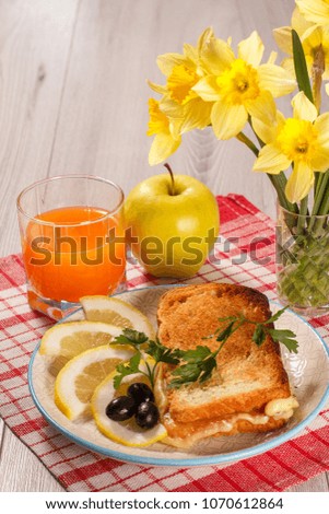 Good and delicious food and beverages for breakfast. Toast with butter and cheese, slices of lemon and olives on plate, apple, bouquet of yellow daffodils and glass of orange juice on kitchen napkin
