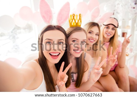 Attractive, charming, cheerful, pretty, stylish girls with crown, bunny ears, eye mask on heads shooting selfie on front camera showing v-sign, having theme party, dress code, sitting on bed