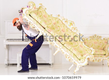Delivery service concept. Loader moves sofa, couch. Man with beard, worker in overalls and helmet carries sofa on back, white background. Courier delivers furniture in case of move out, relocation.
