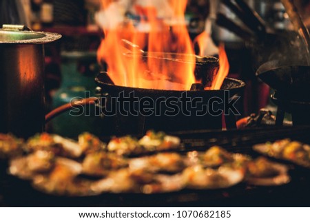 Selective focus picture of some Grilled scallops sold at a street market stall with a pan covered with flames on the background. Chinatown, Bangkok, Thailand