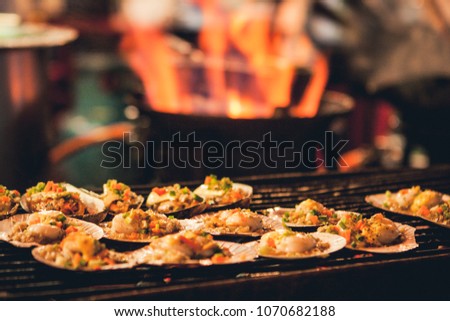 Selective focus picture of some Grilled scallops sold at a street market stall with a pan covered with flames on the background. Chinatown, Bangkok, Thailand