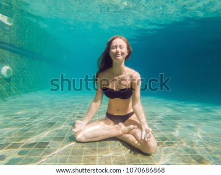 young woman in black bikini in yoga position underwater in diving aquarium, full body shot, front view