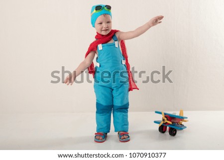 Funny boy in blue overalls and hat, wearing a red scarf and glasses for swimming, sits on vintage suitcase with an airplane model and dreams of adventures. Concept of romance of travel and discovery