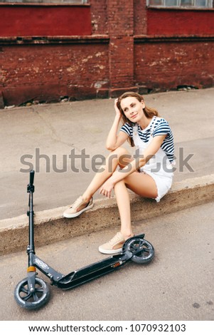 Young beautiful girl sits on a curb in the summer in the city and talks on the phone next to a scooter. The concept of a healthy lifestyle