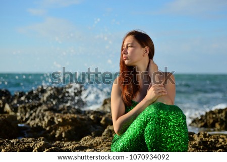 Girl in a shiny costume mermaid sitting on the seashore on the background of the spray and looks at the sunset. Portrait. Horizontal view.