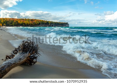 An Upper Peninsula forest creates an autumn background at Chapel Beach in northern Michigan. Lake Superior waves crash on the beach and white puffy clouds dot the blue sky. 