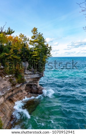 Cumulus clouds dot the horizon as Lake Superior waves crash against a cliff face at Pictured Rocks National Lakeshore, near Munising Michigan. Tree leaves start to show their autumn colors.