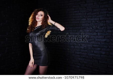 Brunette curly haired long legs girl in black leather dress posed at studio against dark brick wall.
