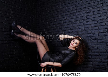 Brunette curly haired long legs girl in black leather dress posed at studio on chair against dark brick wall.