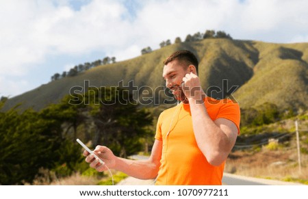 fitness, sport and technology concept - smiling man with smartphone and earphones listening to music over big sur hills and road background in california