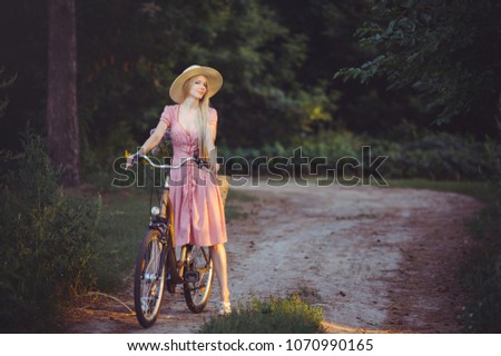 Beautiful girl wearing  nice pink dress having fun in a park with a bicycle holding a beautiful basket with flowers. Vintage landscapes. Pretty blonde with retro look, bicycle and basket with flowers.