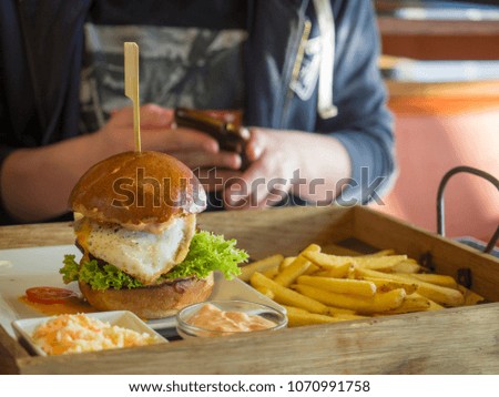 close up big hamburger with fried egg, french fries and barbeque sauce on wooden tray, teenage boy holding cell phone in hands, blurred background