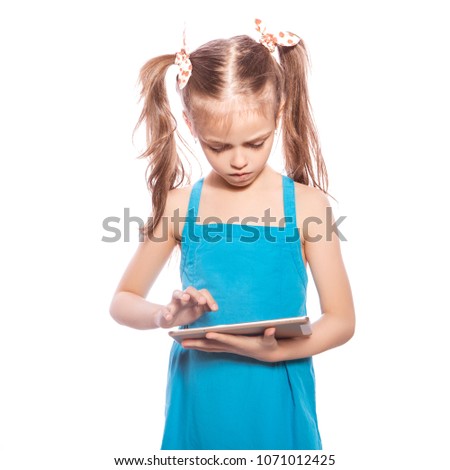 Young seven years old brunette girl in blue dress on a white isolated background. She holds tablet, trying to understand how to use it, thoughtful emotions on her face