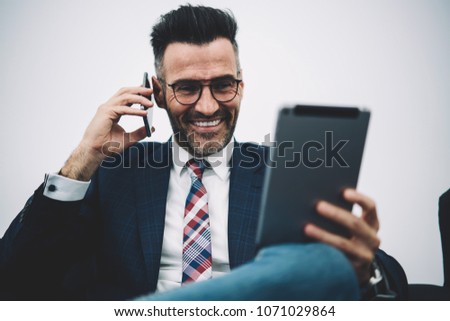 Cheerful mature proud ceo 50 years old dressed in formal wear read funny information on digital touch pad and calling to business partner on smartphone.Happy financial director talking on mobile phone