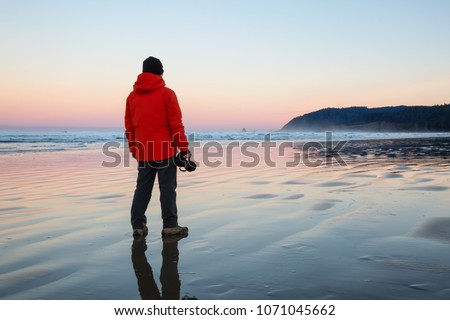 Photographer with a camera is standing on the sandy beach during a vibrant and colorful winter sunrise. Taken in Canon Beach, Oregon Coast, United States of America. Royalty-Free Stock Photo #1071045662