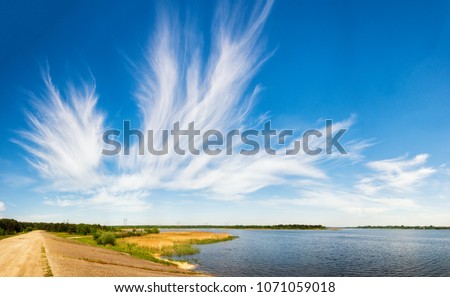 Amazing summer scenery of picturesque lake with reeds on foreground, beautiful cirrus fibratus clouds in the blue sky and dirt road through the dam in countryside. Royalty-Free Stock Photo #1071059018