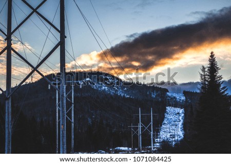 Picture of a mountain covered in snow and tree with a bright cloud from the sunset. Electric cables are running through